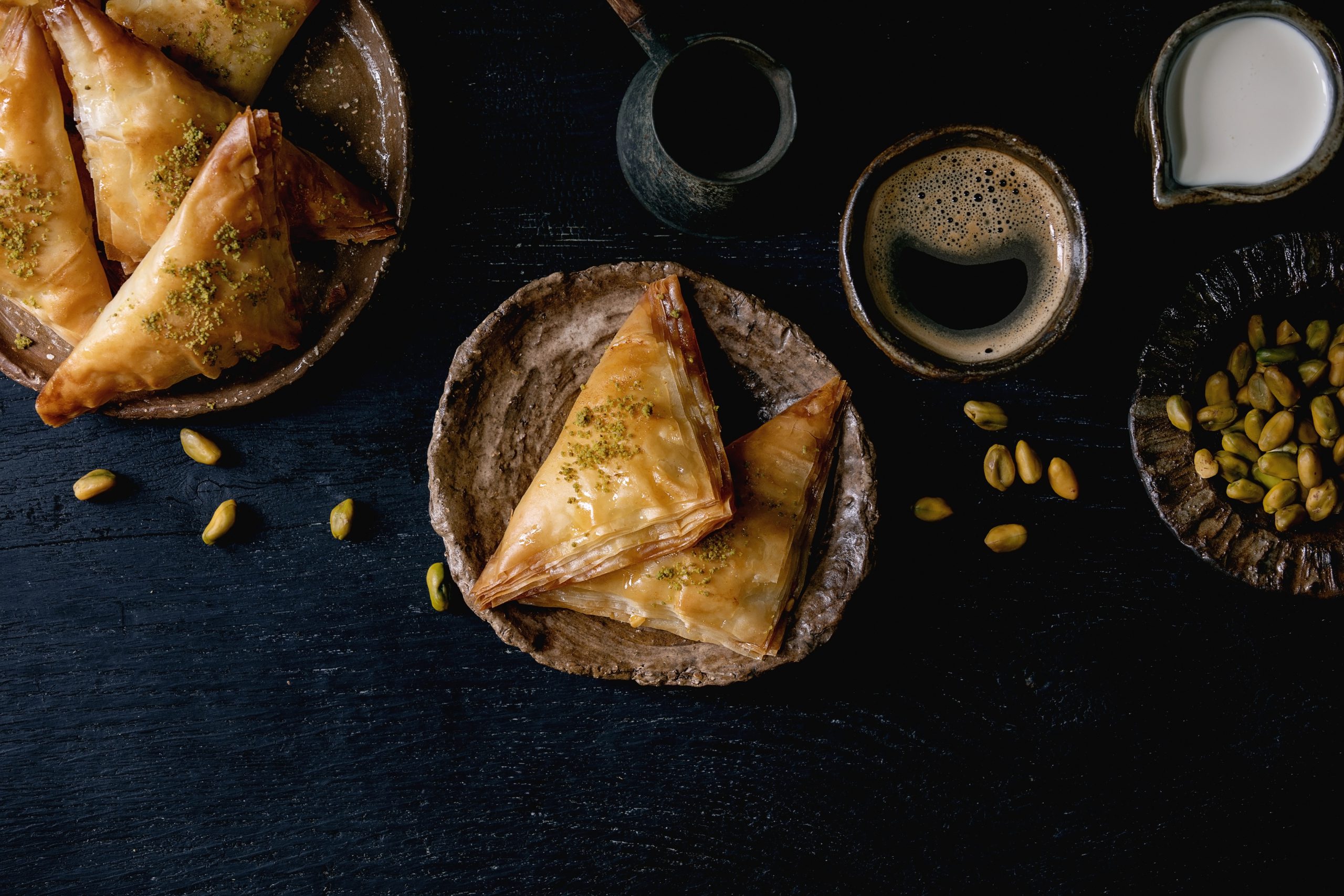 Homemade Turkish traditional dessert baklava with pistachio served on ceramic plate with bowl of nuts, cup of coffee, cream, cezva over black wooden background. Flat lay, space